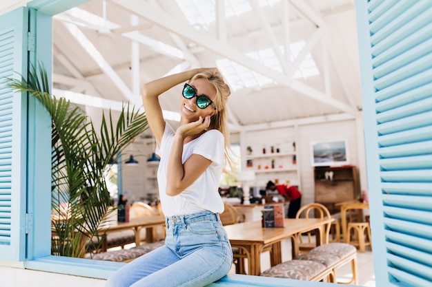 Increíble chica en pantalones casuales vintage sentado en el alféizar de la ventana con una sonrisa. Foto de mujer rubia romántica con gafas brillantes tocando su cabeza.