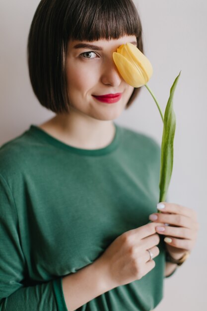 Increíble chica de pelo corto aislada con flor amarilla. Señora caucásica interesada sosteniendo tulipán con linda sonrisa.