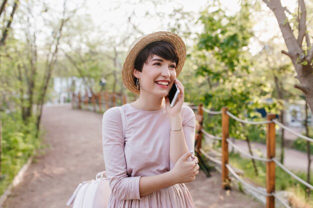 Increíble chica con pelo corto y oscuro hablando por teléfono y mirando hacia arriba con una sonrisa