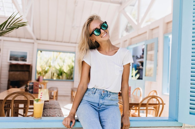 Increíble chica con piel bronceada posando con cóctel de naranja Sonriente mujer rubia con gafas de sol sentada en el alféizar de la ventana en la mañana de verano