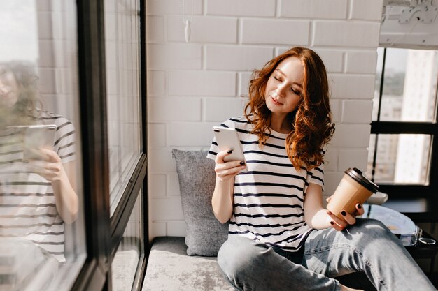 Foto gratuita increíble chica tomando café y mensajes de texto. modelo femenino atractivo que disfruta del café con leche mientras mira la pantalla del teléfono.