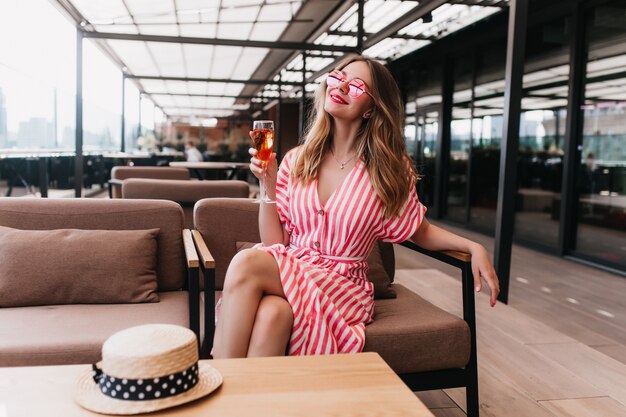 Increíble dama rubia con gafas de sol descansando en la cafetería. Foto interior de hermosa mujer joven en vestido de verano posando con copa de champán en fin de semana.