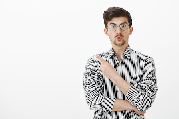 Increible. Hombre europeo interesado sorprendido con barba y bigote en anteojos, doblando los labios y apuntando a la esquina superior izquierda, intrigado y curioso, pidiendo al asistente que muestre un artículo más cercano