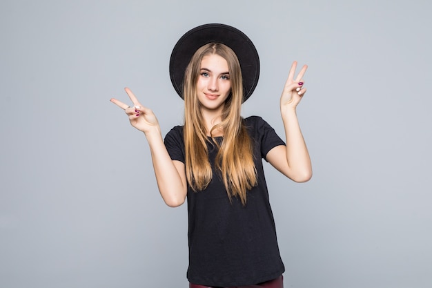 Increíble jovencita con cabello dorado vestida de negro con sombrero retro muestra el signo de la victoria aislado en el fondo