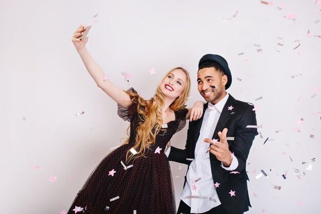 Increíble linda pareja de enamorados celebrando una gran fiesta en oropel, haciendo selfie. Ropa de noche de lujo, emociones positivas brillantes.