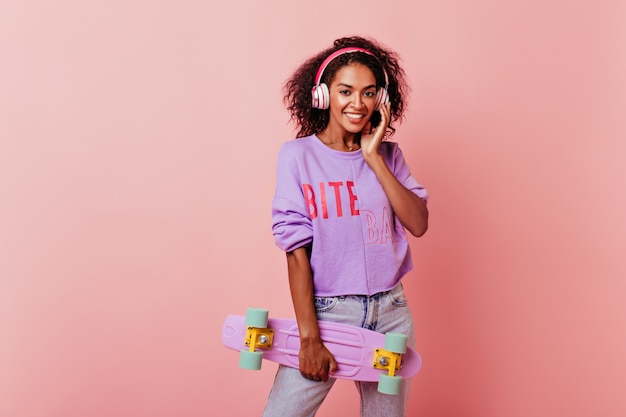 Increíble mujer africana en suéter morado posando con patineta. Debonair modelo femenino negro de pie en rosa en auriculares.