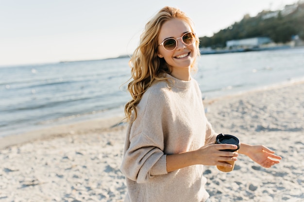 Increíble mujer ciega sosteniendo una taza de café en la playa. Modelo femenino entusiasta en gafas de sol posando cerca del lago en un día frío.