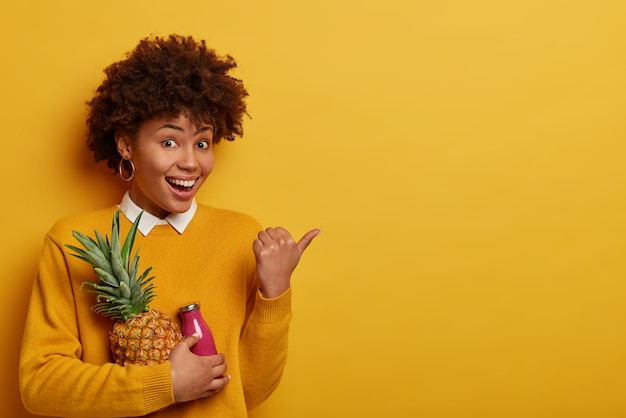 Increíble mujer complacida tiene un estado de ánimo feliz, sostiene piña fresca y una botella de batido, señala con el pulgar a un lado en el espacio de la copia, mantiene la dieta, come frutas que contienen muchas vitaminas, usa ropa amarilla