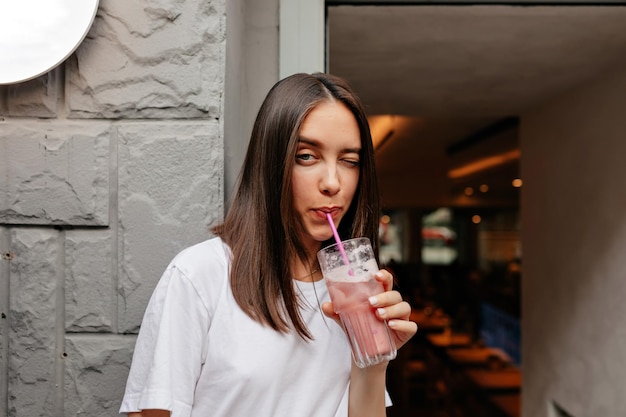 Increíble mujer elegante con cabello oscuro con camiseta blanca posa en la cámara con batido en la ciudad con bebida de verano