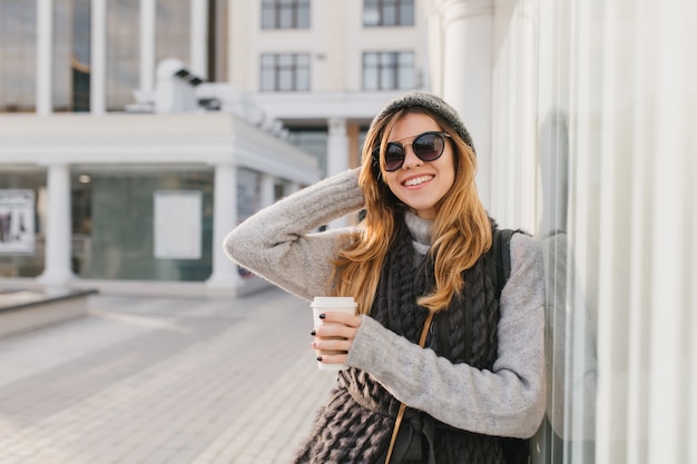 Increíble mujer en elegantes gafas de sol con cabello rubio disfrutando de un café y posando al aire libre con la mano arriba. Retrato de mujer sonriente con sombrero y sudadera tejida de pie en las calles de la ciudad por la mañana.
