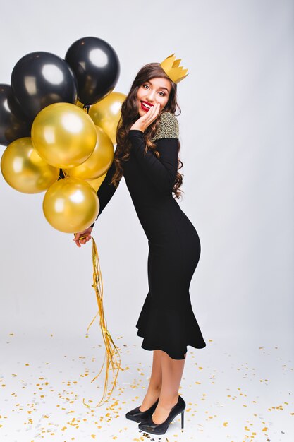 Increíble mujer con estilo alegre en vestido de noche negro de lujo y corona amarilla en la cabeza celebrando el año nuevo, sonriendo y sosteniendo globos amarillos y negros, labios rojos, cara de sorpresa de emoción.