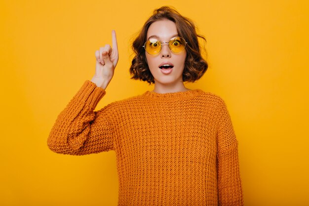 Increíble mujer joven con cabello castaño claro posando con expresión de asombro y levantando el dedo