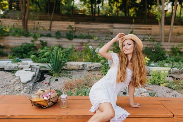 Increíble mujer joven con cabello castaño claro posando mientras toca el sombrero vintage y sonriendo. Hermosa chica en vestido blanco de moda sentado junto a flores con cesta de picnic.