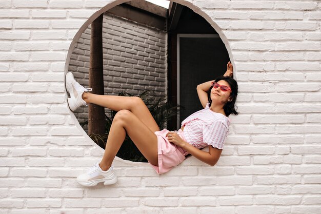 Foto gratuita increíble mujer joven posando en la pared de ladrillos. tiro al aire libre de mujer morena lleva zapatillas blancas.