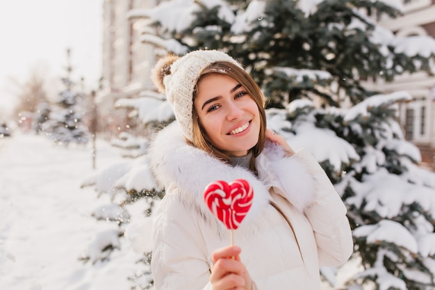 Increíble mujer joven en ropa de abrigo blanca, gorro de punto con lollypop de corazón rosa divirtiéndose en la calle. Mujer atractiva disfrutando de invierno en la ciudad.