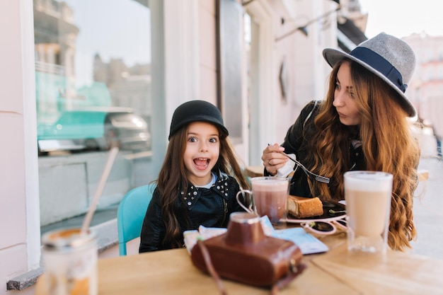 Increíble mujer joven con sombrero gris vintage comiendo sabroso pastel de queso con su hermana pequeña en fin de semana. Chica morena sorprendida disfrutando de un batido de leche en la cafetería al aire libre, mientras que su madre bonita bebe café