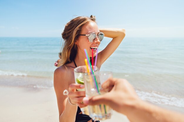 Increíble mujer joven en traje de baño y gafas de sol, bebiendo un cóctel, tostado