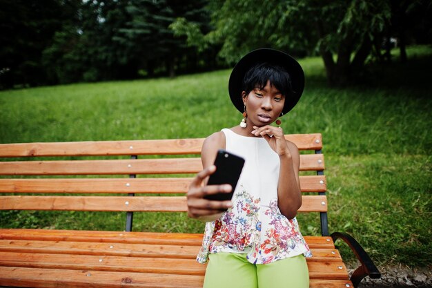 Increíble mujer modelo afroamericana con pantalones verdes y sombrero negro posada en un banco en el parque y haciendo selfie en el teléfono móvil