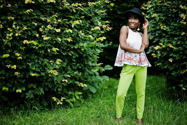 Increíble mujer modelo afroamericana en pantalones verdes y sombrero negro posado en el parque