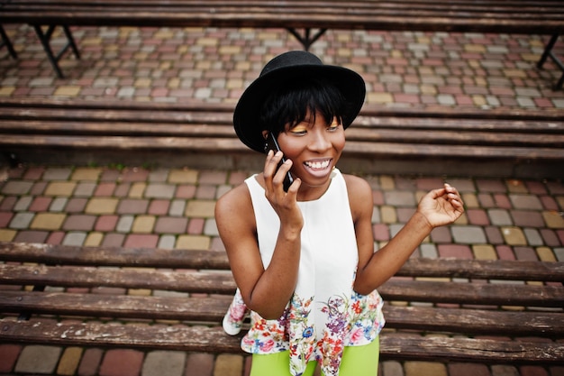 Increíble mujer modelo afroamericana con pantalones verdes y sombrero negro posando en el banco y usando un teléfono inteligente