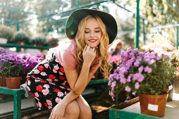 Increíble mujer de pelo largo sentada al lado de flores. Retrato de mujer caucásica entusiasta descansando después de la jardinería.