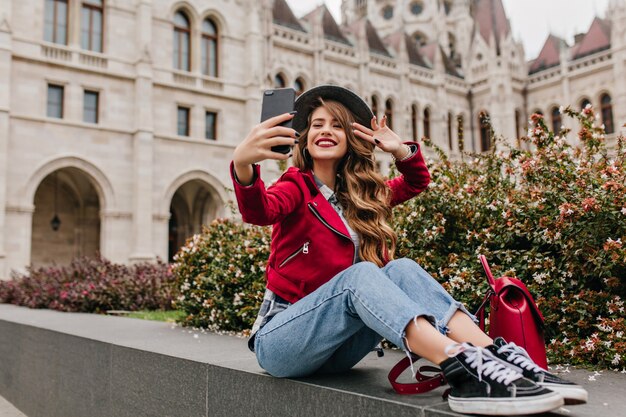 Increíble mujer rizada en zapatos negros haciendo selfie frente al edificio histórico