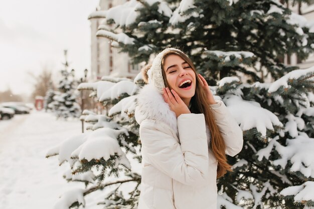 Increíble mujer vestida de blanco divirtiéndose en día de invierno, posando para la foto. Retrato al aire libre de la mujer caucásica complacida en la calle nevada al lado del abeto.