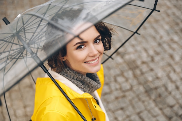 Foto gratuita increíble retrato de mujer joven con abrigo amarillo de pie bajo un paraguas transparente con una amplia sonrisa sincera