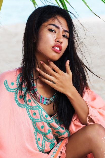 Increíble sexy mujer asiática bronceada posando en la paradisíaca playa tropical bajo el árbol pam, sentada en la arena blanca, relajándose y disfrutando de las vacaciones. Vestido boho con bordado. Bali.