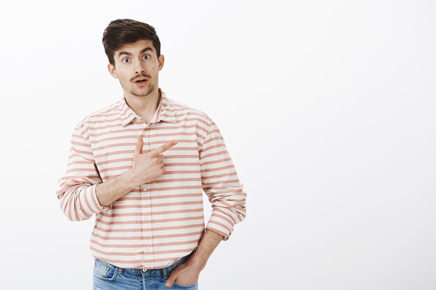 Increíble, tú también lo has visto. Sorprendido novio lindo interesado con bigote y barba, apuntando a la esquina superior derecha con expresión de asombro sobre la pared gris