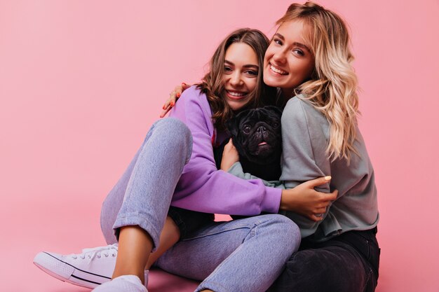 Increíbles hermanas blancas abrazándose durante la sesión de retratos con cachorro. Encantadoras señoritas sentadas en rosa con lindo bulldog.