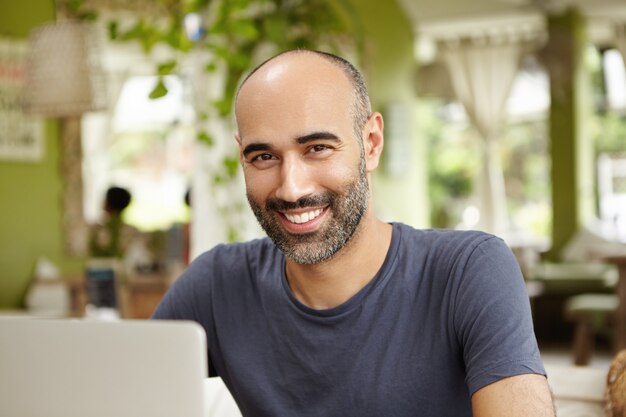 Independiente alegre vestido con una camiseta informal sentado frente a la computadora portátil, mirando y sonriendo con expresión alegre después del exitoso trabajo matutino, disfrutando de un día soleado en un café al aire libre