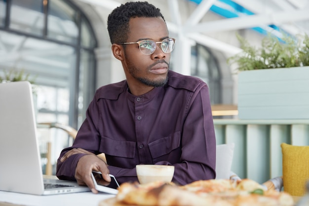 Independiente masculino africano de piel oscura pensativo en elegante camisa, trabaja en un nuevo proyecto, sostiene un teléfono inteligente moderno, bebe café