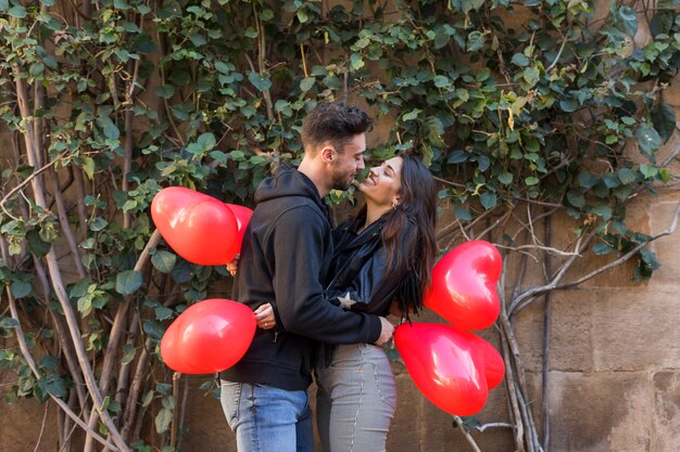 Individuo feliz joven que abraza a la señora sonriente y que sostiene los globos en la forma de corazones