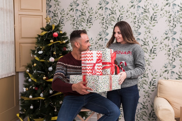 Individuo joven con la pila de regalos y señora atractiva cerca del árbol de navidad