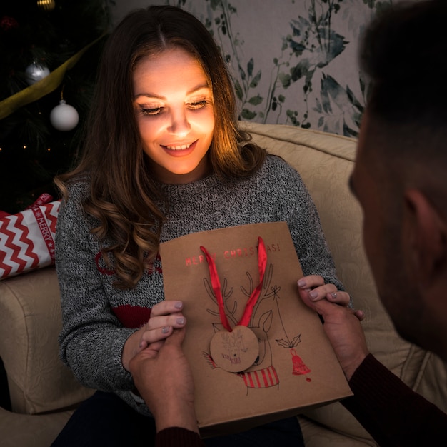 Individuo que presenta el paquete del regalo a la señora atractiva en el settee cerca del árbol de navidad