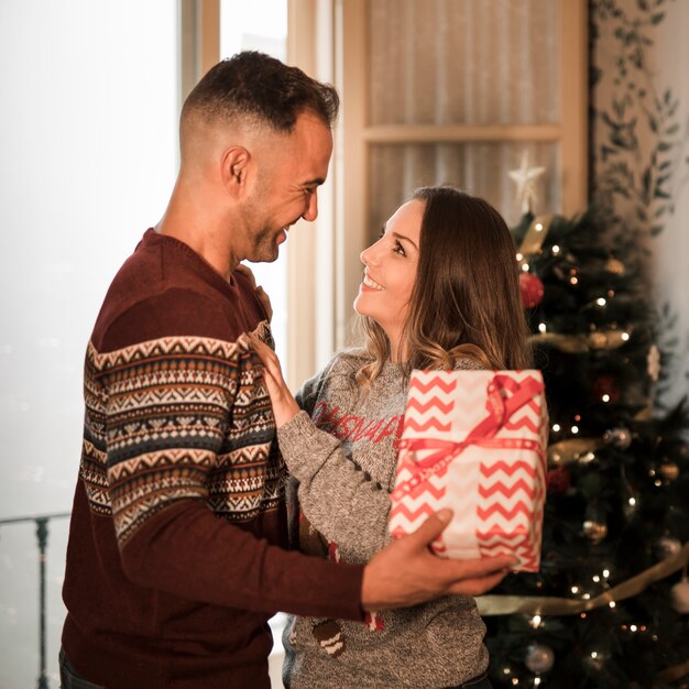 Individuo sonriente con la caja de regalo y la señora alegre cerca del árbol de navidad