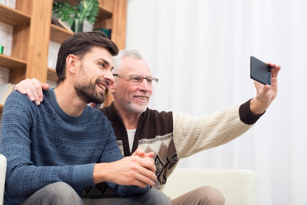 Individuo sonriente joven y hombre alegre envejecido que toman el selfie en el teléfono móvil en el settee