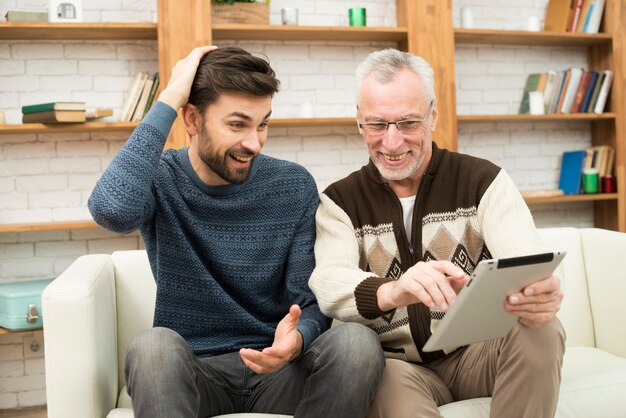 Individuo sonriente joven y hombre alegre envejecido que usa la tableta en el settee