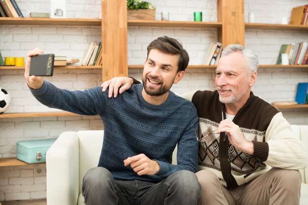 Individuo sonriente joven y hombre envejecido que toman el selfie en smartphone en el settee