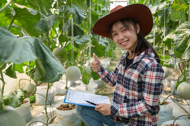 industria agrícola, agricultura, personas y concepto de granja de melón - feliz sonriente mujer joven o agricultor con portapapeles y melón en invernadero granja mostrando pulgares arriba signo de mano