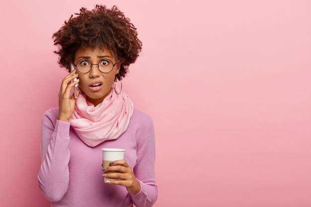 Infeliz, estresada y conmocionada mujer afroamericana habla por teléfono móvil, toma café para llevar, escucha malas noticias, usa anteojos y poloneck púrpura, posa sobre la pared rosada del estudio.