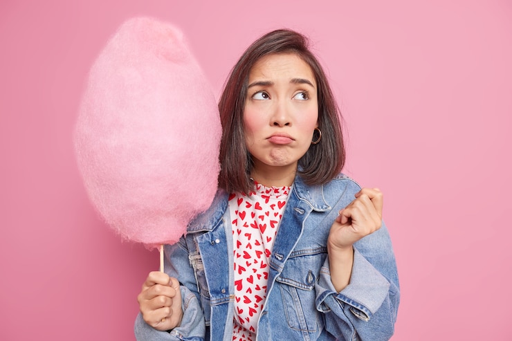 Foto gratuito infeliz frustrada mujer asiática con cabello oscuro mira hacia otro lado tristemente tiene mal humor sostiene algodón de azúcar azucarado mantiene una nutrición saludable no puede romper la dieta usa ropa de mezclilla de moda puestos en interiores