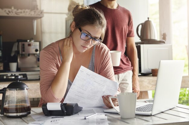 Infeliz hermosa mujer con gafas que tiene mirada concentrada leyendo el formulario de notificación del banco sobre la deuda, sentado en la mesa de la cocina frente a la computadora portátil abierta