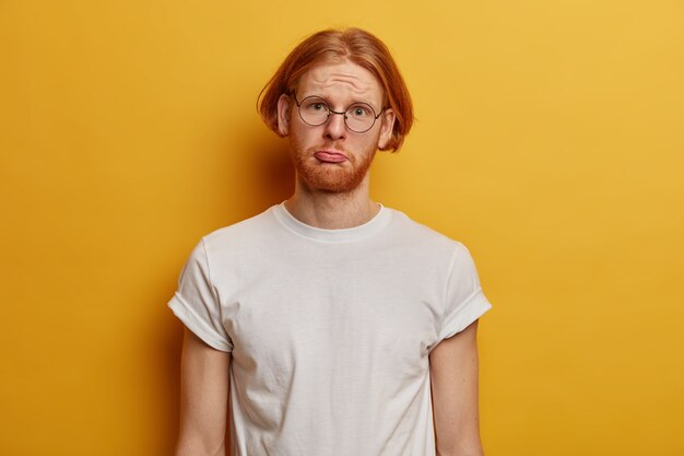 Infeliz hipster chico sombrío con pelo pelirrojo, barba espesa, monederos labio inferior