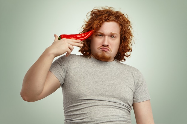 Infeliz hombre joven con sobrepeso obeso y barbudo que se siente miserable y frustrado durante el primer día de su dieta vegetal, atornillando los labios, sosteniendo el pimiento rojo en la sien como si intentara dispararse