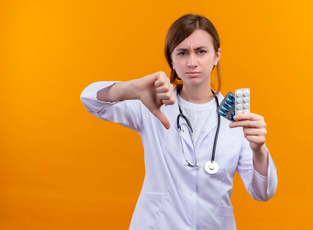 Infeliz joven doctora vistiendo bata médica y estetoscopio sosteniendo medicamentos y mostrando el pulgar hacia abajo en la pared naranja aislada con espacio de copia