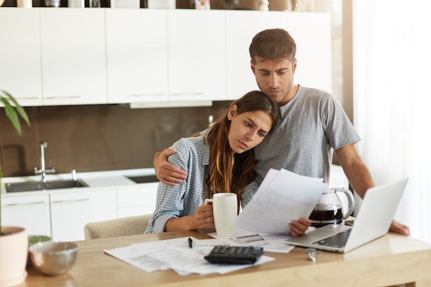 Infeliz joven familia europea que se enfrenta a problemas financieros: triste esposo, sumido en sus pensamientos, abrazando a su preocupada esposa que estudia la notificación del banco en sus manos mientras hace las finanzas en la cocina