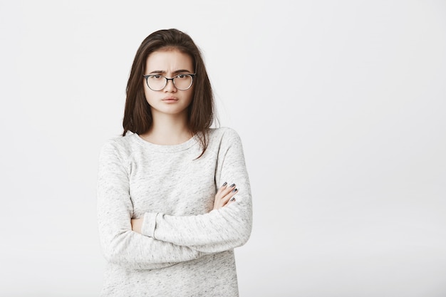 Infeliz joven linda en gafas con expresión sombría y cruza las manos