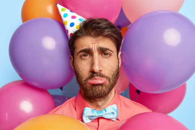 Infeliz joven posando rodeado de globos de colores de cumpleaños